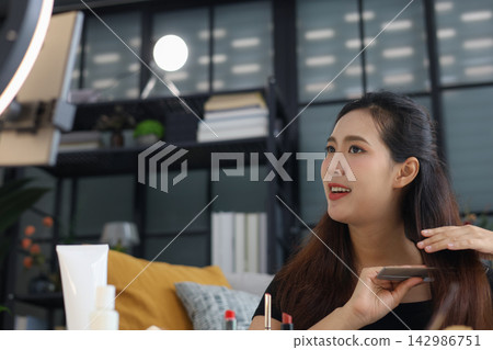 Young Asian woman showing hair tutorial in front of phone camera and ring light. Young Asian woman showing hair tutorial in front of phone camera and ring light. 142986751