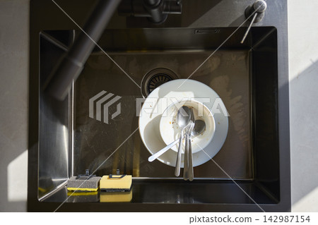 Dirty plates, bowl and cutlery lie in stainless steel kitchen sink beside cleaning sponges Dirty plates, bowl and cutlery lie in stainless steel kitchen sink beside cleaning sponges 142987154