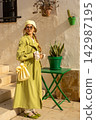 Fashionable woman in green trench coat and beret standing outdoors next to snake plant on sunny day 142987195