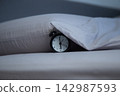 Close-up of a black alarm clock lying on a bed next to a pillow, partially covered by the edge of a gray blanket on a gray background. The concept of getting up in the morning, sleeping patterns, and 142987593