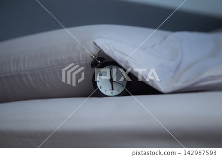 Close-up of a black alarm clock lying on a bed next to a pillow, partially covered by the edge of a gray blanket on a gray background. The concept of getting up in the morning, sleeping patterns, and Close-up of a black alarm clock lying on a bed next to a pillow, partially covered by the edge of a gray blanket on a gray background. The concept of getting up in the morning, sleeping patterns, and 142987593