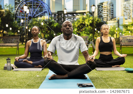 Yoga instructor facing participants seated cross-legged on mats in city park, guiding outdoor meditation and mindful deep breathing session. Trainer and women in Pilates practice. Yoga instructor facing participants seated cross-legged on mats in city park, guiding outdoor meditation and mindful deep breathing session. Trainer and women in Pilates practice. 142987671
