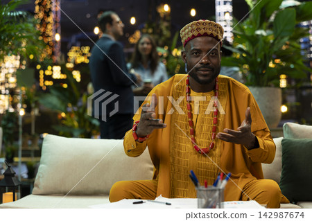 Entrepreneur in traditional attire gestures while speaking in online business teleconference meeting. Businessman looks toward camera and gestures while speaking to remote colleagues from garden Entrepreneur in traditional attire gestures while speaking in online business teleconference meeting. Businessman looks toward camera and gestures while speaking to remote colleagues from garden 142987674