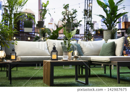 Empty terrace with leafy potted plants and decorative lights placed on small table beside cushioned seating in blurred modern rooftop lounge with scenic ambiance. Empty terrace with leafy potted plants and decorative lights placed on small table beside cushioned seating in blurred modern rooftop lounge with scenic ambiance. 142987675