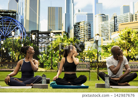 Adults participants cross-legged turning heads while touching necks, performing synchronized mobility stretch during outdoor yoga in city park. Stress relief and flexibility. Adults participants cross-legged turning heads while touching necks, performing synchronized mobility stretch during outdoor yoga in city park. Stress relief and flexibility. 142987676