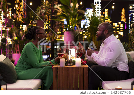 Girlfriend chatting with her man during romantic rooftop date under city lights and candlelight. Boyfriend listens to woman talking, enjoying pleasant conversation over wine Girlfriend chatting with her man during romantic rooftop date under city lights and candlelight. Boyfriend listens to woman talking, enjoying pleasant conversation over wine 142987706