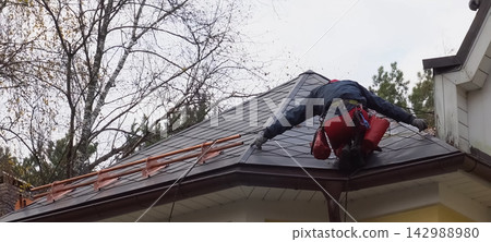 Worker does the installation of the roof of house. Installation of metal tiles, snow retainers and other elements of the roof. Worker does the installation of the roof of house. Installation of metal tiles, snow retainers and other elements of the roof. 142988980
