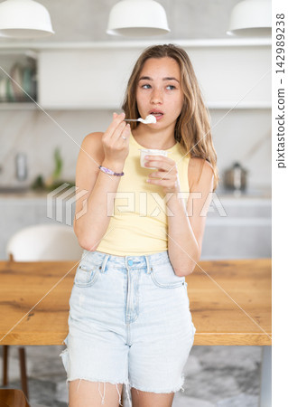 Thoughtful young girl eating fresh yogurt with spoon in modern kitchen Thoughtful young girl eating fresh yogurt with spoon in modern kitchen 142989238