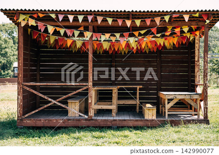 Cozy autumn outdoor wooden pavilion decorated with orange and yellow handmade bunting flags featuring rustic timber furniture on a green grass lawn. Cozy autumn outdoor wooden pavilion decorated with orange and yellow handmade bunting flags featuring rustic timber furniture on a green grass lawn. 142990077