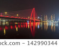 A red illuminated cable-stayed bridge spans a river beside a modern city skyline, with colorful lights reflecting across the water at night. 142990344