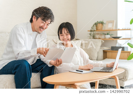 A high school girl is getting tutoring from her dad at home. Father and daughter studying together while looking at textbooks. A high school girl is getting tutoring from her dad at home. Father and daughter studying together while looking at textbooks. 142990702