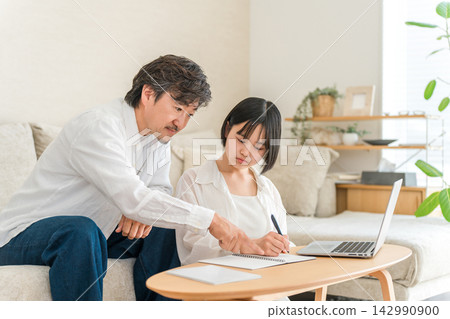A high school girl is being tutored by her father at home. Father and daughter studying using notebooks. A high school girl is being tutored by her father at home. Father and daughter studying using notebooks. 142990900