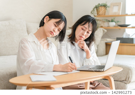 A high school daughter is tutored by her education-focused mother. The mother and daughter feel pressured by the need for home study. A high school daughter is tutored by her education-focused mother. The mother and daughter feel pressured by the need for home study. 142991273