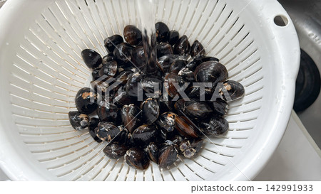The clams are placed in a white colander and washed under running water before cooking. The clams are placed in a white colander and washed under running water before cooking. 142991933