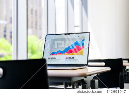 A laptop computer sits on a desk by the window, displaying a graph. A laptop computer sits on a desk by the window, displaying a graph. 142991947