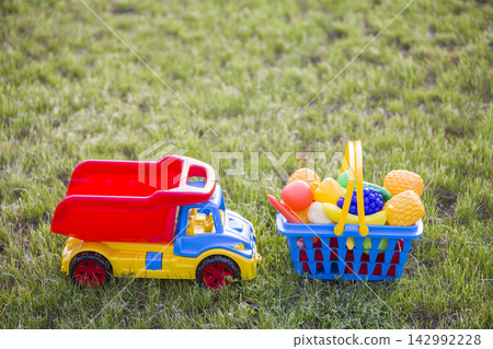 Car truckand a basket with toy fruit and vegetables. Bright plastic colorful toys for children outdoors on sunny summer day. Car truckand a basket with toy fruit and vegetables. Bright plastic colorful toys for children outdoors on sunny summer day. 142992228