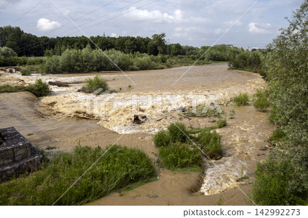 Fast flowing brown river overflowing its banks after torrential rain, illustrating flood damage, erosion, climate change and destructive force of nature. Fast flowing brown river overflowing its banks after torrential rain, illustrating flood damage, erosion, climate change and destructive force of nature. 142992273