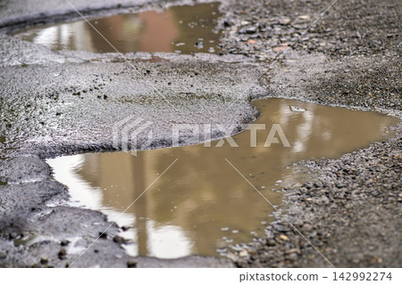 Dilapidated city street surface with broken asphalt and water filled potholes. Infrastructure decline, unsafe travel and urban maintenance issues. Dilapidated city street surface with broken asphalt and water filled potholes. Infrastructure decline, unsafe travel and urban maintenance issues. 142992274