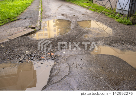 Large potholes filled with dirty rainwater on city street, creating a dangerous obstacle for cars while illustrating road decay and public infrastructure problems. Large potholes filled with dirty rainwater on city street, creating a dangerous obstacle for cars while illustrating road decay and public infrastructure problems. 142992276