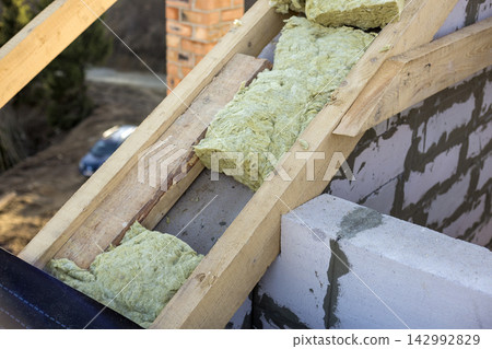 Roof construction and insulation with mineral wool. Wooden beams frame on walls of hollow foam insulation blocks. Roofing underlayment, water-resistant waterproof barrier material as protection. Roof construction and insulation with mineral wool. Wooden beams frame on walls of hollow foam insulation blocks. Roofing underlayment, water-resistant waterproof barrier material as protection. 142992829