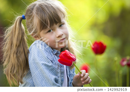 Cute pretty smiling child girl with gray eyes and long hair with bright red tulip flower on blurred sunny green bokeh background. Love to nature concept. Cute pretty smiling child girl with gray eyes and long hair with bright red tulip flower on blurred sunny green bokeh background. Love to nature concept. 142992919