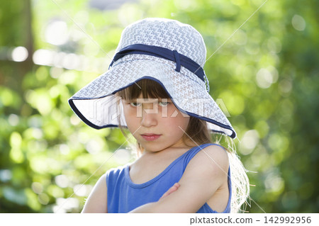 Close-up portrait of serious little girl in a big hat. Child having fun time outdoors in summer. Close-up portrait of serious little girl in a big hat. Child having fun time outdoors in summer. 142992956