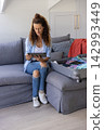 Mature woman sitting on gray sofa at home holding tablet and packing suitcase with passport 142993449