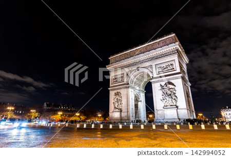 Low angle night view of the illuminated triumphal arch monument Arc de Triomphe with street cobblestones and night sky in Paris, France. Low angle night view of the illuminated triumphal arch monument Arc de Triomphe with street cobblestones and night sky in Paris, France. 142994052