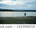 Back portrait of woman stands alone in front of the lake with horizontal line of forest and mountain in background 142994056