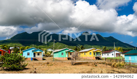 Row of vibrant colorful wooden houses in a rural Caribbean village with lush green mountains and cloudy blue sky in Saint Kitts and Nevis. Row of vibrant colorful wooden houses in a rural Caribbean village with lush green mountains and cloudy blue sky in Saint Kitts and Nevis. 142994062
