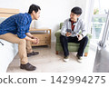 Asian men sitting and talking in bright bedroom, one holding smartphone and small booklet 142994275