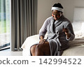 African American adult woman sitting on bed in robe with under-eye patches applying from dropper 142994522