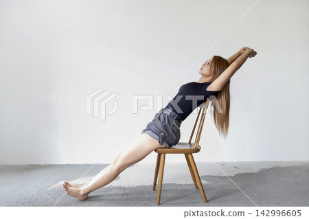 A woman stretches while sitting in a chair in a minimalist and simple studio. 142996605