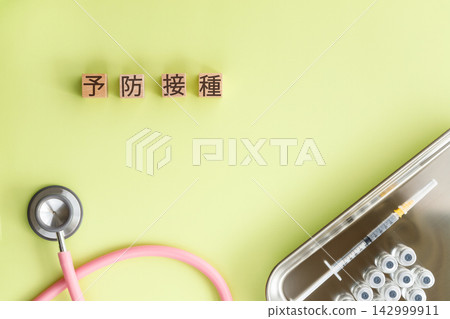 Wooden cube with the word "vaccination" written on it, along with a syringe and a stethoscope, against a light green background. Wooden cube with the word "vaccination" written on it, along with a syringe and a stethoscope, against a light green background. 142999911