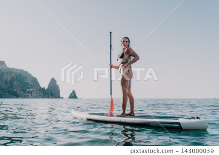 Woman Paddleboarding Ocean Mountains - A woman in a bikini stands on a paddleboard in the ocean with mountains in the background. Woman Paddleboarding Ocean Mountains - A woman in a bikini stands on a paddleboard in the ocean with mountains in the background. 143000039