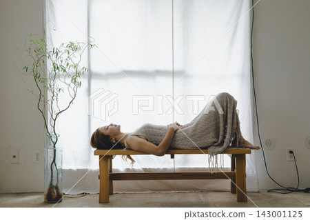 一位美麗的日本女子在沐浴著自然光的窗邊長椅上放鬆地冥想。 一位美麗的日本女子在沐浴著自然光的窗邊長椅上放鬆地冥想。 143001125