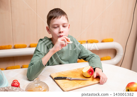 A young boy eating a slice of a red apple at a table with a wooden cutting board and a kitchen knife in front of him A young boy eating a slice of a red apple at a table with a wooden cutting board and a kitchen knife in front of him 143001382