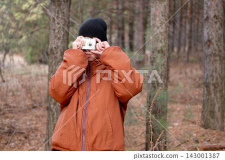 preteen boy in an orange jacket and black beanie holding a silver compact camera in front of his face while taking a picture in a pine forest preteen boy in an orange jacket and black beanie holding a silver compact camera in front of his face while taking a picture in a pine forest 143001387