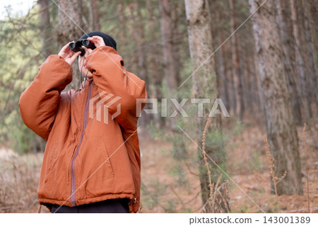 A young boy wearing an orange jacket and black knit beanie holding binoculars to his eyes and looking up towards trees in a pine forest A young boy wearing an orange jacket and black knit beanie holding binoculars to his eyes and looking up towards trees in a pine forest 143001389