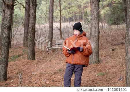 young preteen boy wearing an orange jacket, gloves, and a knit beanie reading a book while standing in a pine forest young preteen boy wearing an orange jacket, gloves, and a knit beanie reading a book while standing in a pine forest 143001391
