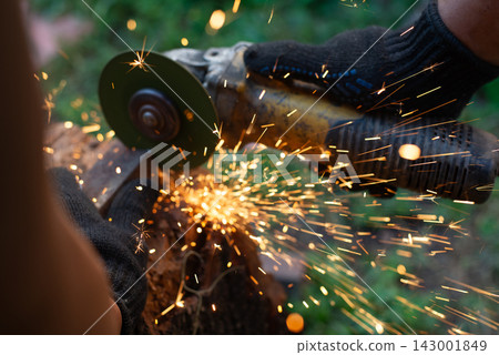 Man in protective gloves cutting metal with an angle grinder outdoors. Motion blur bright sparks flying. Concept of manual labour, home workshop, DIY metalwork and work safety. Man in protective gloves cutting metal with an angle grinder outdoors. Motion blur bright sparks flying. Concept of manual labour, home workshop, DIY metalwork and work safety. 143001849
