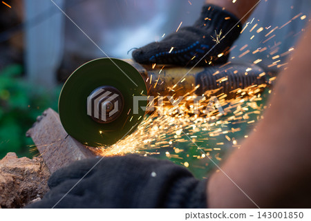 Man in protective gloves cutting metal with an angle grinder outdoors, bright sparks flying. Concept of manual labour, home workshop, DIY metalwork and work safety. Man in protective gloves cutting metal with an angle grinder outdoors, bright sparks flying. Concept of manual labour, home workshop, DIY metalwork and work safety. 143001850