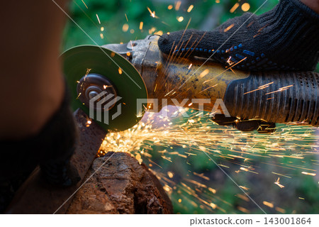 Man in protective gloves cutting metal with an angle grinder outdoors, bright sparks flying. Concept of manual labour, home workshop, DIY metalwork and work safety. 143001864