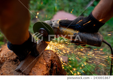 Man in protective gloves cutting metal with an angle grinder outdoors, bright sparks flying. Concept of manual labour, home workshop, DIY metalwork and work safety. Man in protective gloves cutting metal with an angle grinder outdoors, bright sparks flying. Concept of manual labour, home workshop, DIY metalwork and work safety. 143001867