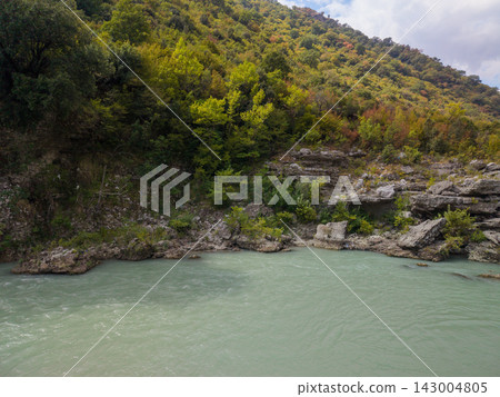 Vjosa River Albania flows past rocky bank below forested slope. Green water and rugged limestone create wild riverscape, with dense woodland rising under soft daylight in tranquil mountain setting. Vjosa River Albania flows past rocky bank below forested slope. Green water and rugged limestone create wild riverscape, with dense woodland rising under soft daylight in tranquil mountain setting. 143004805