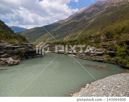 Vjosa River Albania runs between rugged limestone banks and tree covered slopes, with still water in foreground. Vast alpine scenery creates quiet, dramatic mood in Vjosa River Albania. Vjosa River Albania runs between rugged limestone banks and tree covered slopes, with still water in foreground. Vast alpine scenery creates quiet, dramatic mood in Vjosa River Albania. 143004806