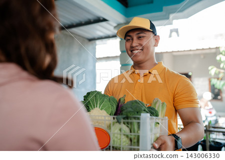 Smiling produce seller in orange polo and cap hands fresh vegetables from basket to customer in bright indoor market with soft daylight and friendly service atmosphere Smiling produce seller in orange polo and cap hands fresh vegetables from basket to customer in bright indoor market with soft daylight and friendly service atmosphere 143006330