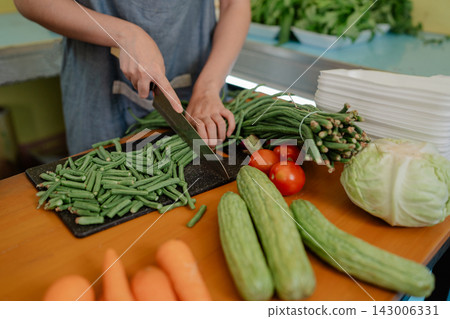 Person slicing fresh green beans beside tomatoes, cucumbers, cabbage and carrots in bright kitchen prep scene with healthy vegetables, cutting board and knife for meal preparation Person slicing fresh green beans beside tomatoes, cucumbers, cabbage and carrots in bright kitchen prep scene with healthy vegetables, cutting board and knife for meal preparation 143006331