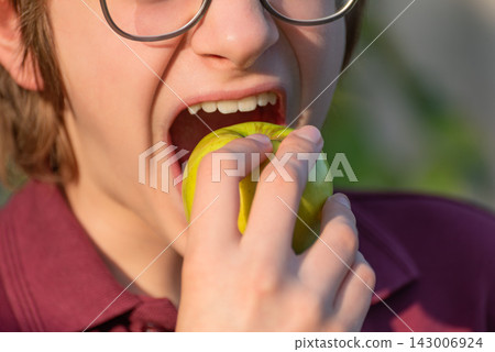 Young person with glasses opening mouth wide to bite a crisp green apple, close up promoting healthy eating, fresh fruit snack, nutrition and enjoyment of natural, crunchy food Young person with glasses opening mouth wide to bite a crisp green apple, close up promoting healthy eating, fresh fruit snack, nutrition and enjoyment of natural, crunchy food 143006924