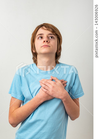 Young boy standing with hands placed over chest, looking upward with a thoughtful and sincere expression, reflecting concepts of hope, gratitude, and spirituality 143006926